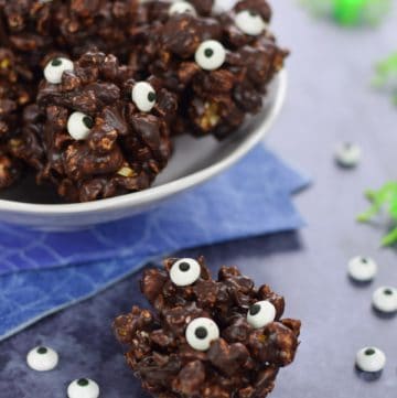 Single monster popcorn ball with a bowl of popcorn balls behind and scattered candy eyeballs